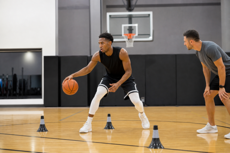 Basketball player performing a controlled court drill while a trainer observes in an indoor gym